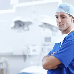 Chirurg w niebieskim stroju i czepku, ze skrzyżowanymi ramionami, na tle sali operacyjnej.