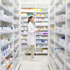 Farmaceutka w białym fartuchu idzie między regałami pełnymi leków, szukając milurit 300.