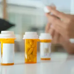 Leki w pomarańczowych buteleczkach na stole, w tle ręce przygotowujące kolejną dawkę. Na horyzoncie nadzieja na poprawę.