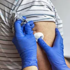 Ręce w niebieskich rękawiczkach wykonują iniekcję w ramię pacjenta. Obok igły widać wacik.