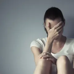 Kobieta w białej koszulce siedzi przy szarej ścianie, zakrywając twarz dłońmi. Czuć w niej **deprexolet** – smutek i przygnębienie.