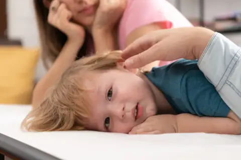 Małe dziecko z blond włosami leży na białym materacu, jego mama patrzy z troską. Czy to początek pandas choroba?