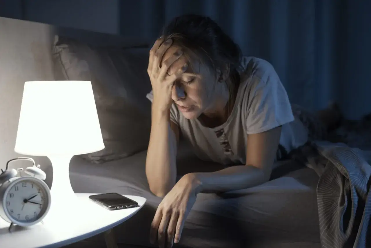 Kobieta w łóżku, z ręką na czole, zmaga się z bezsennością. Co to jest insomnia? Zegar i telefon obok sugerują bezowocne próby zaśnięcia.