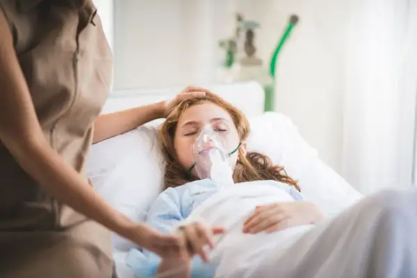 Dziewczynka w szpitalnym łóżku z maską tlenową, walcząca z hipoksją. Opiekunka czule kładzie dłoń na jej głowie.